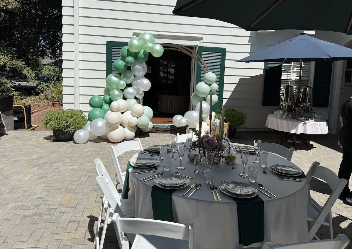 Sage and white balloon arch over a doorway with fresh stems worked into the base beside a styled table — balloon and floral styling for Watsonville catering