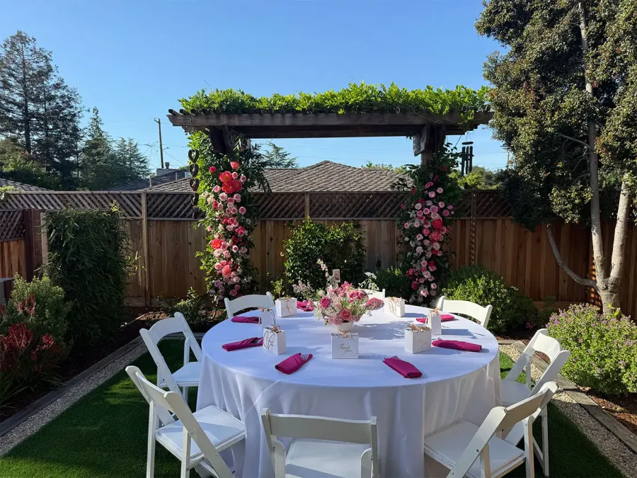 Rose arch in blush and deep red rising above a round garden table with white linen and fuchsia napkins — ceremony arch florals designed alongside Santa Cruz catering